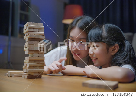 Smiling woman help teach child play build constructor of wooden blocks, Asian young mother playing game in wood block with her little daughter in home living room at night time before going to bed 123198348