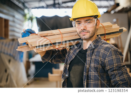 Smiling construction worker in a yellow hardhat and plaid shirt holding wood planks on his shoulder. Ideal for carpentry, woodworking, and craftsmanship in furniture workshop, National Carpenters Day Smiling construction worker in a yellow hardhat and plaid shirt holding wood planks on his shoulder. Ideal for carpentry, woodworking, and craftsmanship in furniture workshop, National Carpenters Day 123198354