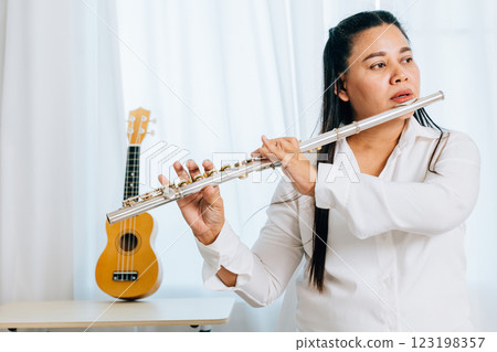 A woman in her living room sits on a chair, dressed in a white shirt, playing a silver flute. The flowing curtains in the background frame her in a moment of musical expression. 123198357