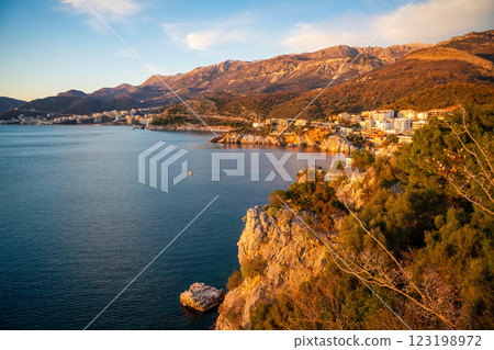 Coastal view of Becici and Budva from a rocky cliff, bathed in golden sunset light with the sea stretching toward the horizon. 123198972