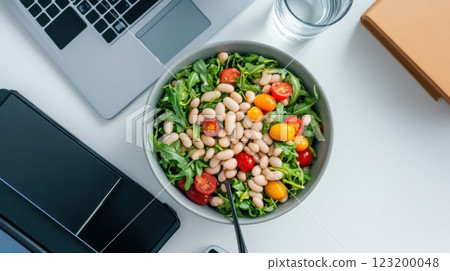 A healthy office lunch with bean salad, white beans, arugula, and cherry tomatoes, served on a desk beside a laptop and water glass. A healthy office lunch with bean salad, white beans, arugula, and cherry tomatoes, served on a desk beside a laptop and water glass. 123200048