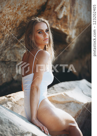 Elegant blonde woman in a white swimsuit sitting gracefully on a rock, blending fashion and nature. 123202405