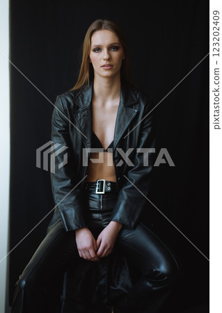 Powerful sophistication concept - woman in black leather, seated with an intense gaze, her presence commanding the scene, as the dark backdrop and polished styling enhance the high-fashion mood 123202409