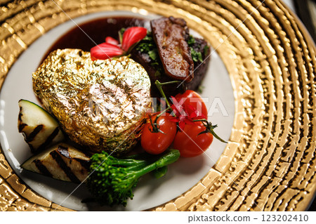 Opulent gold-plated beef steak with grilled vegetables, cherry tomatoes, and a flavorful sauce. Adorned with edible flowers, served on a chic plate in a luxurious restaurant setting 123202410