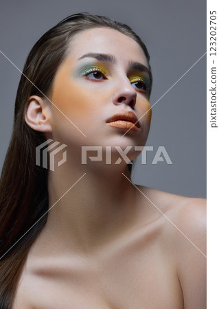 Portrait of beautiful young woman gazing upwards with golden-toned lips and vibrant green eyeshadow against gray background. 123202705