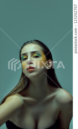 Young woman tilting head slightly while wearing bright green and yellow artistic makeup against teal background. Young woman tilting head slightly while wearing bright green and yellow artistic makeup against teal background. 123202707
