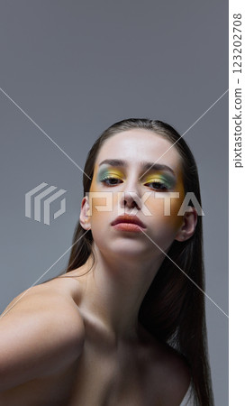 Artistic portrait of young, beautiful woman with green and yellow artistic makeup gazing confidently against soft gray background. Artistic portrait of young, beautiful woman with green and yellow artistic makeup gazing confidently against soft gray background. 123202708