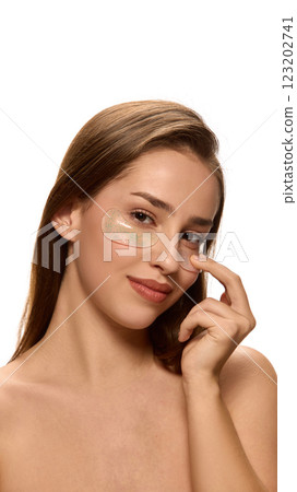 Young woman gently pressing under-eye patches, cosmetic product, while smiling, looking at camera against white background. Young woman gently pressing under-eye patches, cosmetic product, while smiling, looking at camera against white background. 123202741