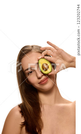 Young woman holding avocado half over eye with soft smile against white background, representing organic cosmetology products. Young woman holding avocado half over eye with soft smile against white background, representing organic cosmetology products. 123202744