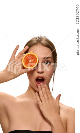 Young woman holding fresh orange slice over eye, and looking at camera with surprised expression against white background. Young woman holding fresh orange slice over eye, and looking at camera with surprised expression against white background. 123202746