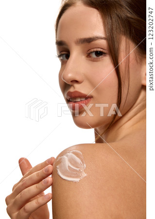 Young woman applying moisturizing cream, beauty product on shoulder and looking at camera against white background. 123202747