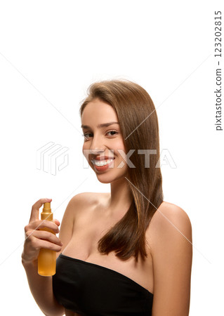 Young woman in black stylish top holding hair spray while looking at camera and smiling against white studio background. 123202815