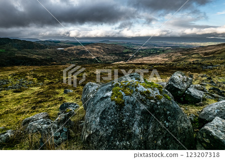 Dark grey rocks of varying sizes are piled on a hillside. A view of a broad, flat valley under a cloudy sky extends beyond the hill. Dark grey rocks of varying sizes are piled on a hillside. A view of a broad, flat valley under a cloudy sky extends beyond the hill. 123207318