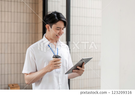 Young Asian male office worker standing and smiling while using tablet in modern workspace, enjoying coffee and digital interaction. 123209316