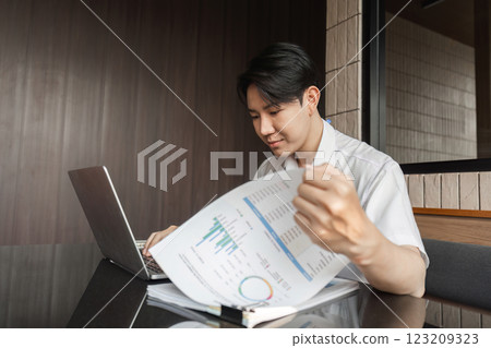 Young professional analyzing financial report while working on laptop in modern office environment. Young professional analyzing financial report while working on laptop in modern office environment. 123209323