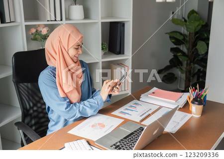 Muslim woman smiling while using smartphone in a modern office with financial documents. Muslim woman smiling while using smartphone in a modern office with financial documents. 123209336