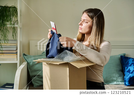 Serious upset young woman unpacking box with online purchases, looking at clothes 123210521