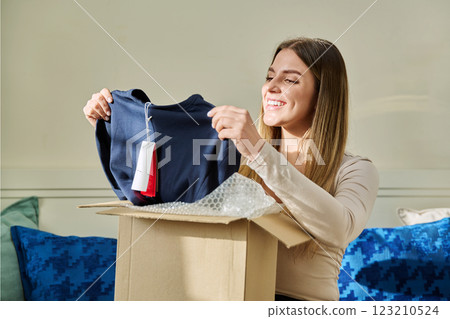 Young woman unpacking cardboard box parcel with online order, looking at new clothes Young woman unpacking cardboard box parcel with online order, looking at new clothes 123210524