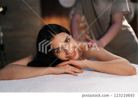 Serene woman relaxing during back massage in spa with soft light, smiling looking at camera. Beautiful naked female with perfect skin getting relaxing massage. Concept of rest with spa treatment. 123210693