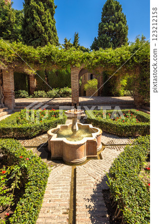 Alcazaba, palace and fortification in Malaga city at Andalusia, Spain, Europe. Serene courtyard garden with a stone fountain, meticulously manicured hedges, and brick pathways. A tranquil oasis. 123212158