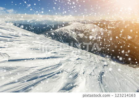 Beautiful winter landscape. Steep mountain hill slope with white deep snow, distant mountain range panorama, large snowflakes and bright shining sun rays on blue sky colorful copy space background. Beautiful winter landscape. Steep mountain hill slope with white deep snow, distant mountain range panorama, large snowflakes and bright shining sun rays on blue sky colorful copy space background. 123214163