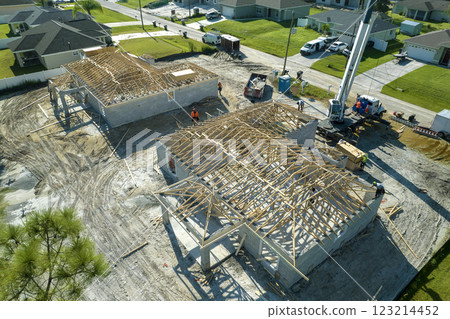 Aerial view of lifting crane and builders working on unfinished residential house with wooden roof frame structure under construction in Florida suburban area 123214452