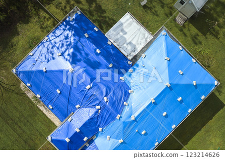 Aerial view of damaged in hurricane Ian house roof covered with blue protective tarp against rain water leaking until replacement of asphalt shingles Aerial view of damaged in hurricane Ian house roof covered with blue protective tarp against rain water leaking until replacement of asphalt shingles 123214626