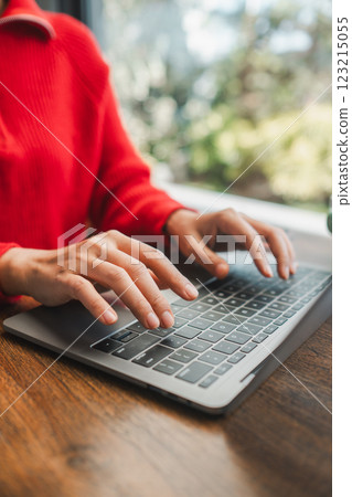 Hands typing on a laptop keyboard, wearing a red sweater, in a cozy environment with natural light. 123215055
