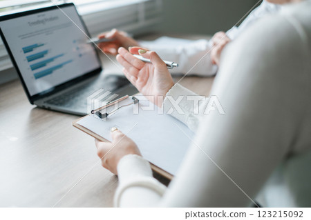 Professionals reviewing financial charts on a laptop, discussing strategy with clipboard and pen in a modern office environment. 123215092