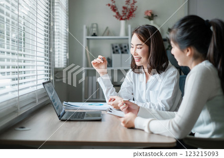Two professional women working together at a desk, discussing business strategies with a laptop and documents in a modern office setting. 123215093