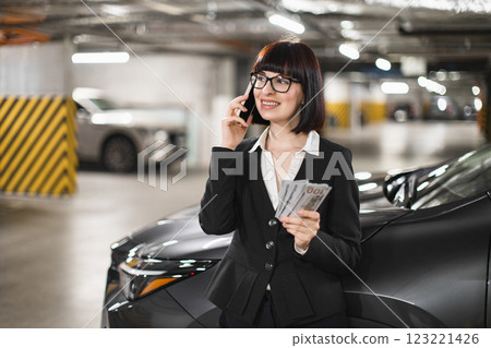 Caucasian businesswoman in parking lot holding money while on phone call. Professional appearance with confident demeanor. Image reflects themes of finance business and communication in urban setting. 123221426
