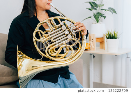 A female musician sits on her sofa at home, blowing a gleaming French horn. The cozy living room setting complements the elegance of her gold instrument. A female musician sits on her sofa at home, blowing a gleaming French horn. The cozy living room setting complements the elegance of her gold instrument. 123222709