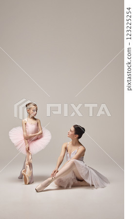 Young girl in pink tutu holding pointe shoes, standing confidently while adult ballerina sits nearby observing with smile against light background. Young girl in pink tutu holding pointe shoes, standing confidently while adult ballerina sits nearby observing with smile against light background. 123225254