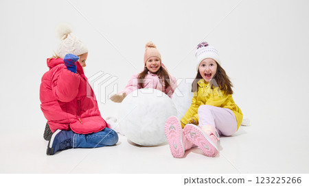 Three girls sitting near large snowball, laughing and preparing for next step in building snowman against white studio background. Three girls sitting near large snowball, laughing and preparing for next step in building snowman against white studio background. 123225266