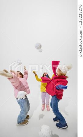 Happy, little girls playing with snowballs, throwing them into air with joy, wearing colorful winter jackets against white studio background. 123225276