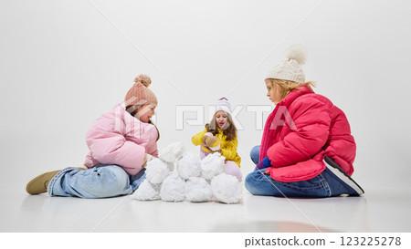Three girls in colorful winter outfits sitting on floor, rolling snowballs and having fun together against white studio background. Three girls in colorful winter outfits sitting on floor, rolling snowballs and having fun together against white studio background. 123225278