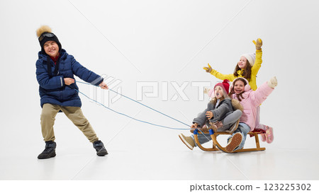 Boy pulling sled with three girls, friends, sitting, all dressed in cozy winter outfits, enjoying playful ride against white studio background. Boy pulling sled with three girls, friends, sitting, all dressed in cozy winter outfits, enjoying playful ride against white studio background. 123225302