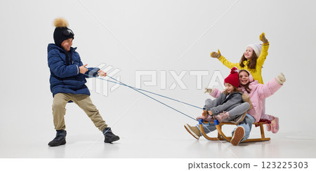 Boy pulling sled with three girls sitting, all dressed in cozy winter outfits, enjoying playful ride against white studio background. Boy pulling sled with three girls sitting, all dressed in cozy winter outfits, enjoying playful ride against white studio background. 123225303