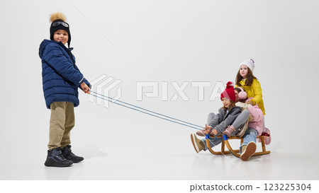 Boy pulling sled with friends sitting, dressed in warm winter coat, hat with pompom, showing effort against white studio background. Boy pulling sled with friends sitting, dressed in warm winter coat, hat with pompom, showing effort against white studio background. 123225304