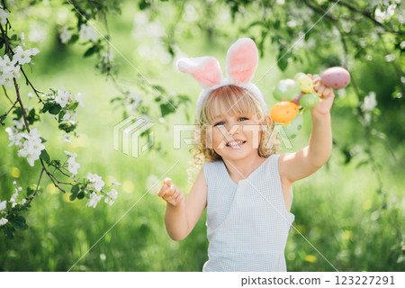 Easter egg hunt. Girl child Wearing Bunny Ears Running To Pick Up Egg In Garden. Easter tradition. Baby with basket full of colorful eggs. 123227291