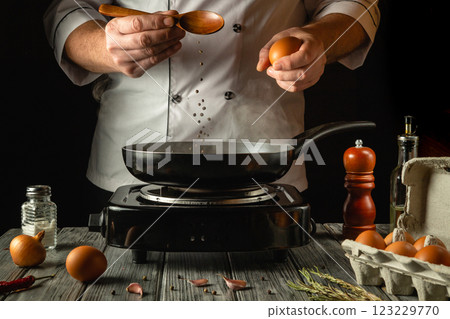 A skilled chef expertly holds an egg above a hot frying pan and spoon, surrounded by ingredients like spices and herbs on a rustic wooden table. The atmosphere hints at culinary creativity and passion 123229770