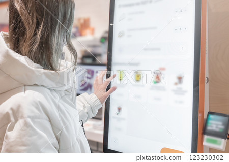 A woman in a white jacket chooses drinks and orders food for dessert in a restaurant or pizzeria coffee shop through a touchscreen screen on her own. The restaurant menu is displayed on the screen in 123230302