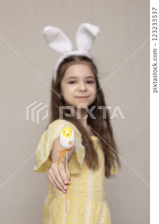 Girl in yellow dress holding Easter-themed egg decoration wearing bunny ear headband 123233537