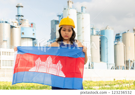 Sad young woman in helmet holding flag of Cambodia against background of factory Sad young woman in helmet holding flag of Cambodia against background of factory 123236532