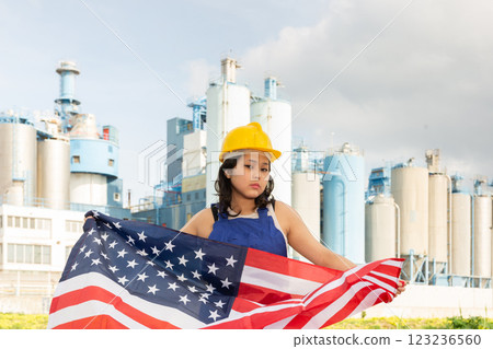 Portrait of upset girl in construction overalls with the USA flag on the background of modern factory 123236560