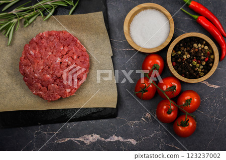 The concept of cooking a burger, fast food, natural products. Close-up left of beef raw round patty lying on paper next to seasonings, herbs and vegetables on dark background. High quality photo 123237002