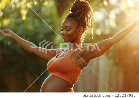 Pregnant woman in orange top with arms outstretched, standing in sunny garden. Concept of joy, peace, and motherhood. For maternity and lifestyle content. 123237985