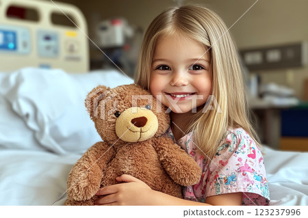 Young girl in a hospital bed smiling and hugging a toy bear, wearing colorful pajamas, with medical equipment blurred in the background. 123237996
