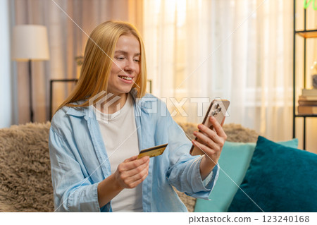 Young woman paying with credit card, smartphone transaction, relief, success at home couch, sunset 123240168