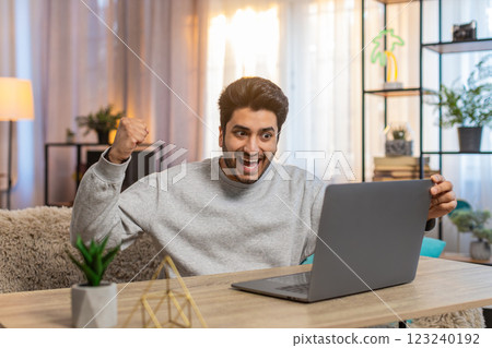 Young Indian man using laptop celebrating after receiving important email and good news at table 123240192